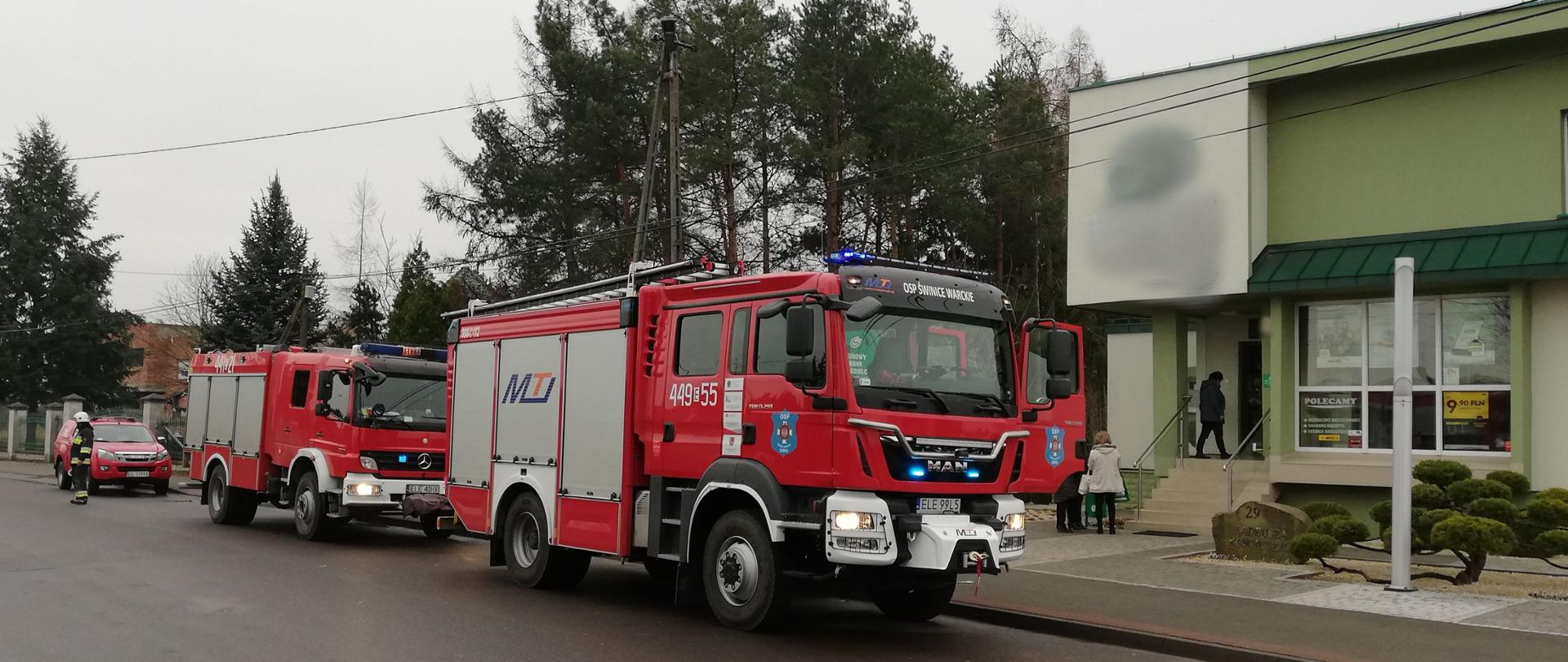 Na zdjęciu widać trzy czerwone samochody strażackie stojące na ulicy przed budynkiem banku, w którym doszło do pożaru. Na jezdni stoi strażak w ciemnym ubraniu i białym hełmie, który kieruje ruchem. W tle widać osoby stojące przed budynkiem banku. Jest widno.