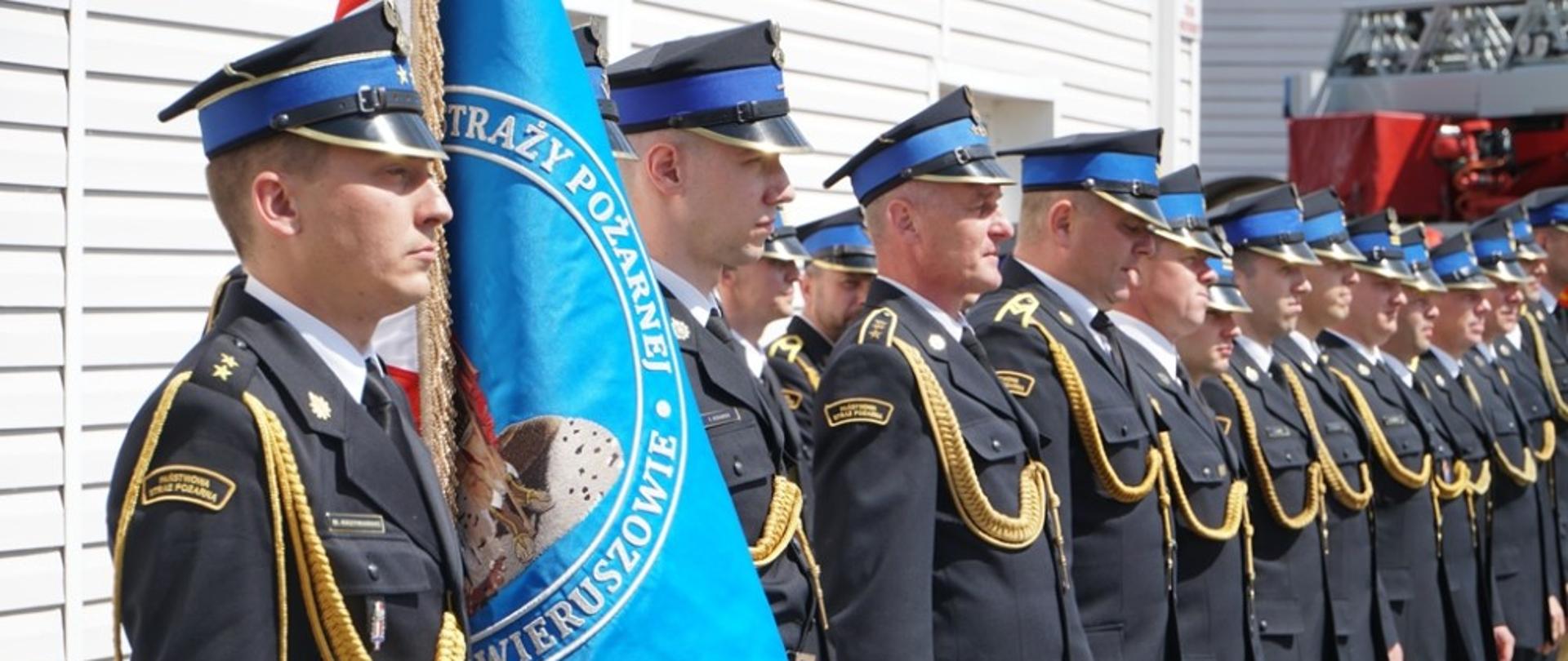 Zdjęcie przedstawia strażaków Państwowej Straży Pożarnej w umundurowaniu wyjściowym stojących w dwuszeregu. na pierwszym planie widać sztandar jednostki