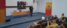 Mężczyzna trzyma mikrofon za nim ustawione są urządzenia fotowoltaiczne obok stoi baner Komendy Wojewódzkiej PSP w Gdańsku na ścianie jest logo miasta Słupsk przed mężczyzną w rzędach na fotelach siedzą strażacy.
