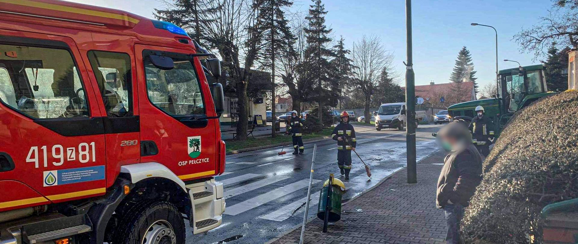 Na pierwszym planie zdjęcia po lewej stronie znajduję się samochód pożarniczy przed samochodem jest dwóch strażaków. Obaj mają w rękach miotłę i usuwają z drogi elementy po kolizji. Po prawej stronie na chodniku stoi widz i przygląda się pracy strażaków. W tle widać poruszające się samochody osobowe oraz ciągnik rolniczy stojący na poboczu, który uczestniczył w zdarzeniu.