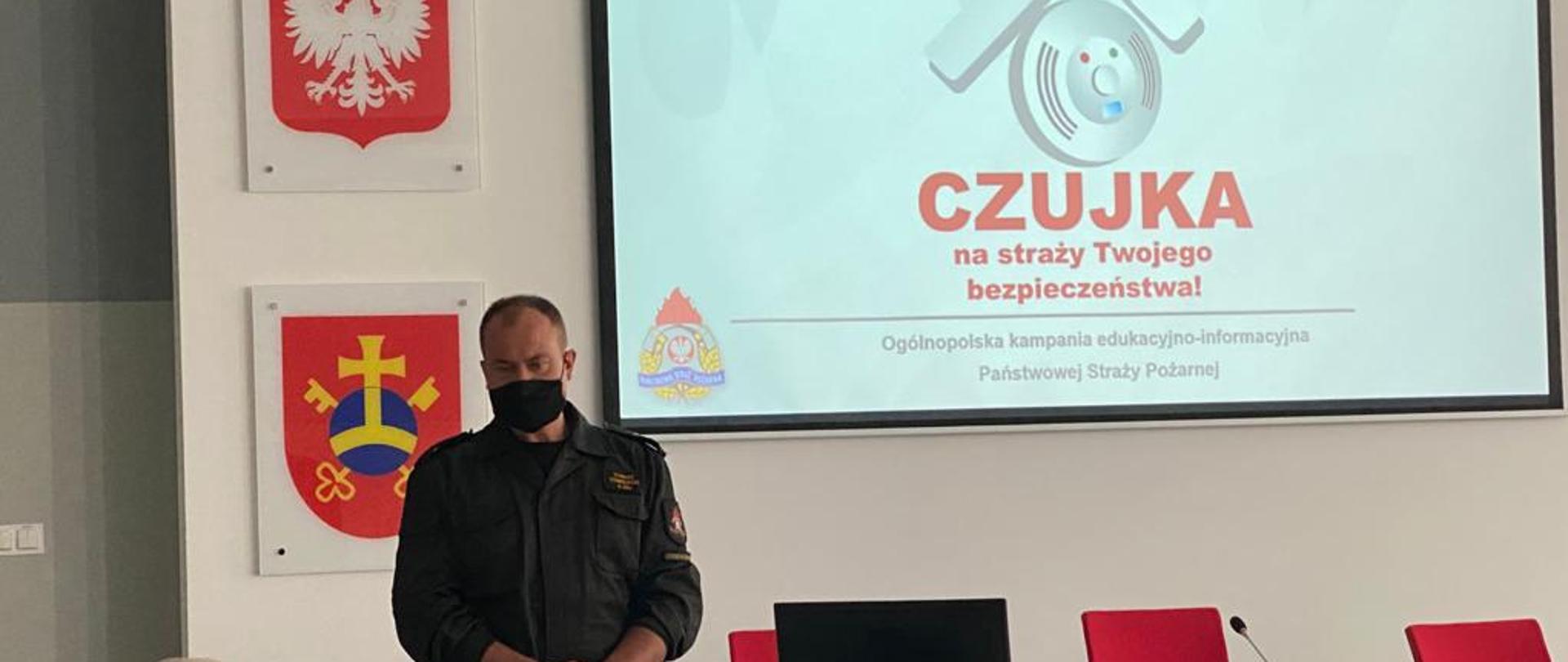 Funkcjonariusz opowiada seniorom o bezpieczeństwie pożarowym. Przed nim widoczny jest jeden senior. Za Funkcjonariuszem znajduje się ekran wyświetlający prezentację, a obok niego godło i herb miasta Ostrowa Wielkopolskiego.