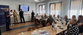 Na zdjęciu znajdują się strażak, policjant oraz kilka osób siedzących za ławkami a w tle widać baner policji oraz ekran z wyświetloną treścią