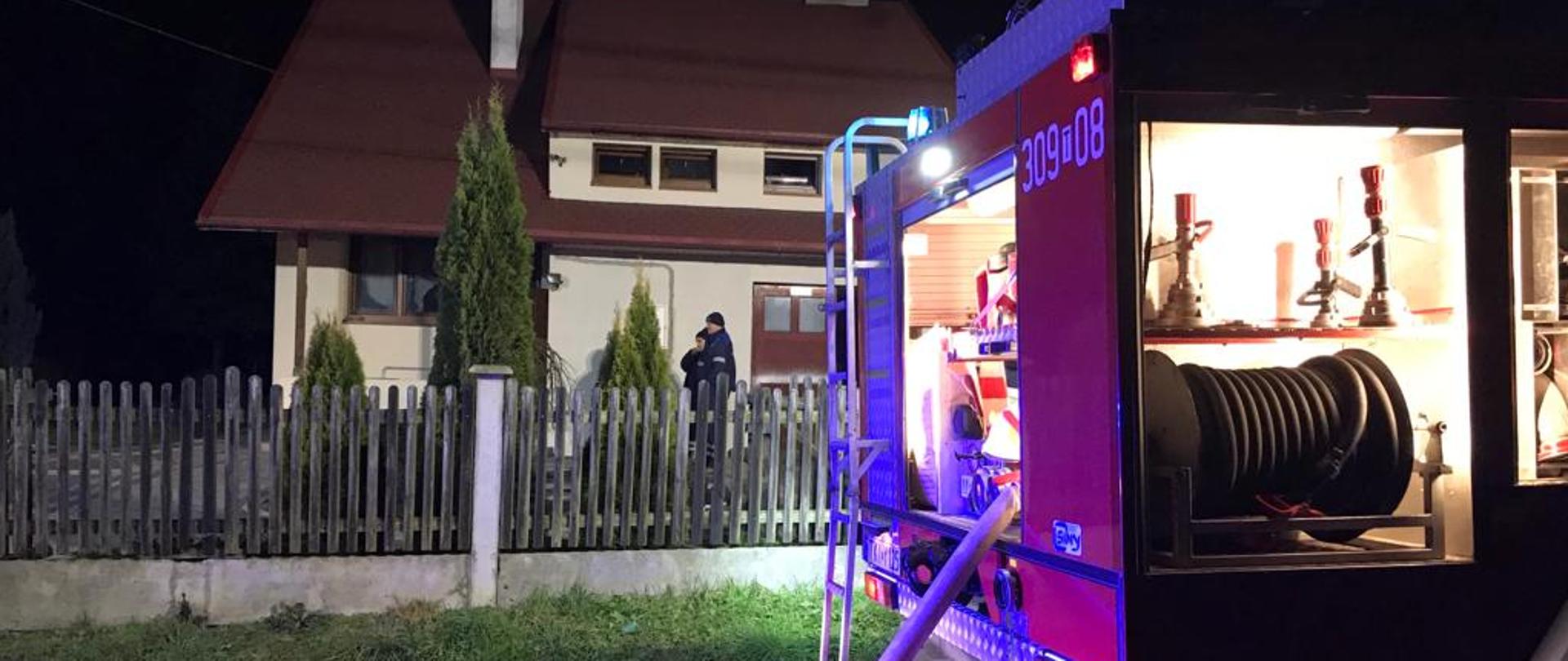 Zdjęcie przedstawia zaparkowany samochód pożarniczy z podpiętym wężem gaśniczym. W głębi, za ogrodzeniem widać dom mieszkalny, w którym wybuchł pożar.