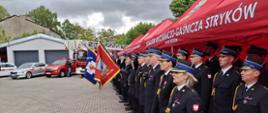 Zdjęcie przedstawia pododdziały strażaków oraz poczty sztandarowe PSP i OSP z powiatu Zgierskiego, w tle widać pojazdy pożarnicze. 