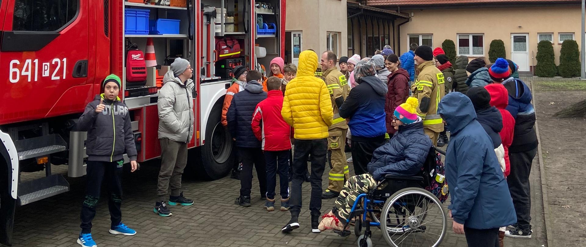 Na pierwszym planie dzieci wraz z strażakami oglądały samochód pożarniczy. Po lewej stronie średni wóz strażacki, natomiast w tle budynek zespołu szkół specjalnych w Wolsztynie. 