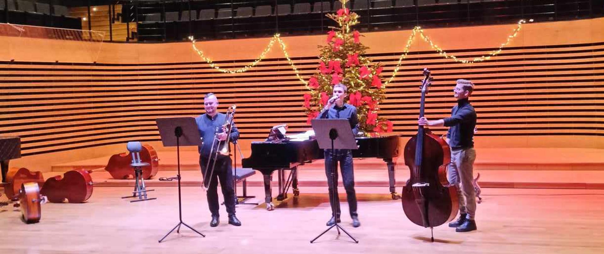 Trzech mężczyzn stoi na estradzie sali koncertowej, jeden trzyma puzon, drugi mikrofon, trzeci kontrabas. Za nimi stoi fortepian.