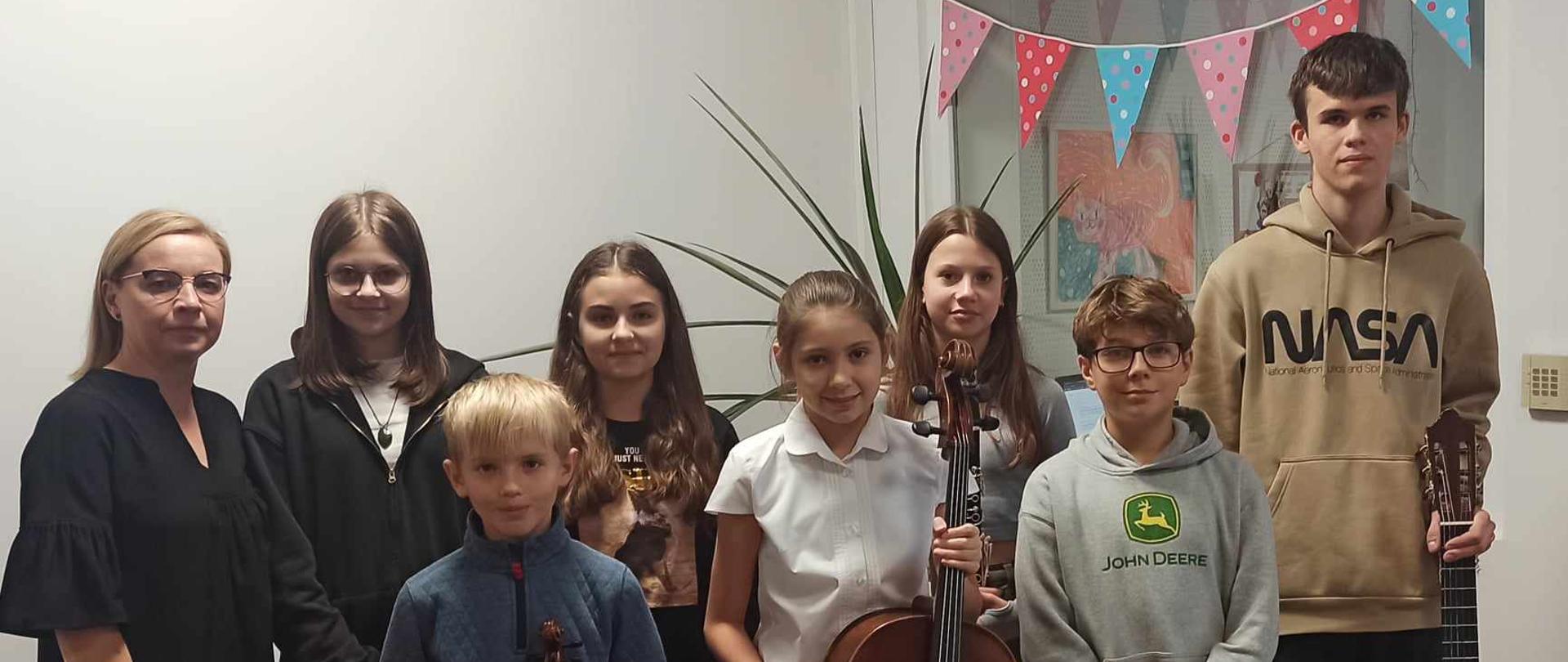 Zdjęcie przedstawia siedmioro uczniów w pozycji stojącej, wraz z nauczycielką. Uczniowie pozują do zdjęcia trzymając instrumenty muzyczne.