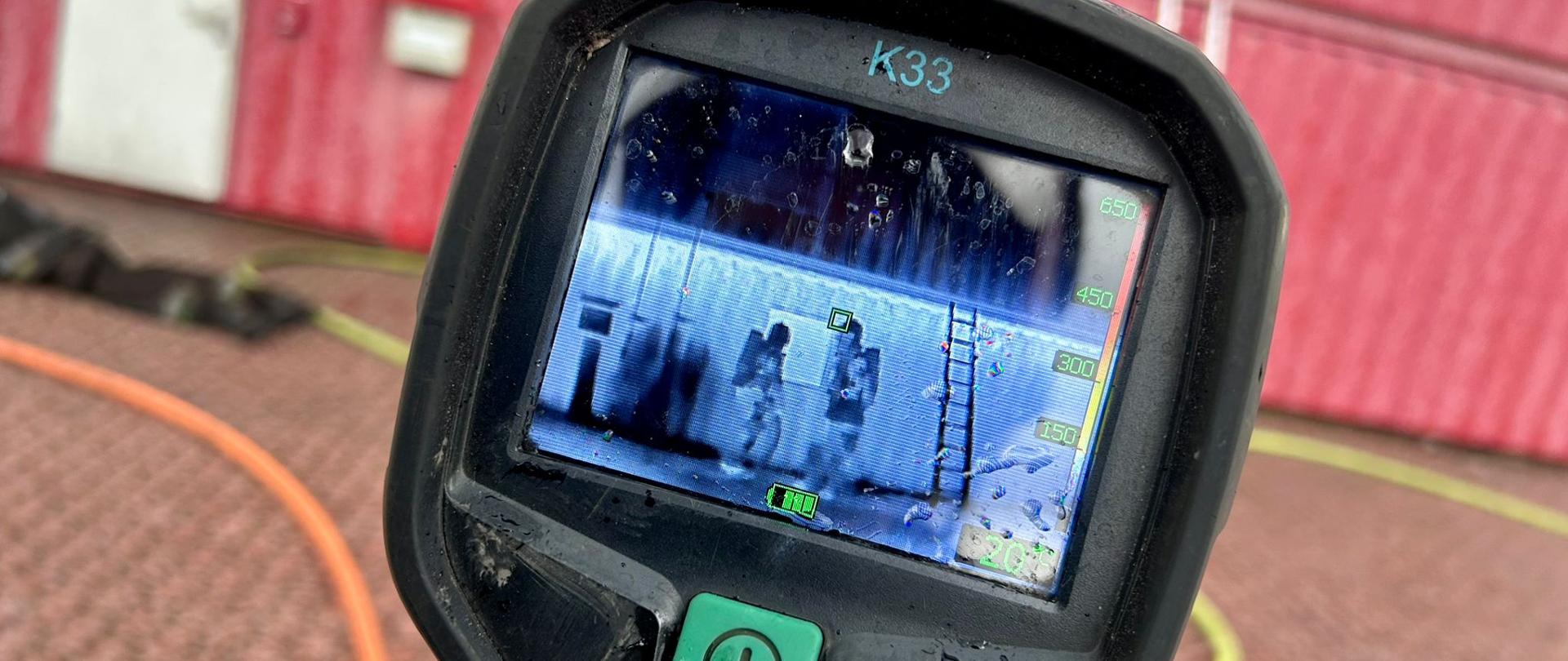 Zbliżenie na ekran kamery termowizyjnej pokazujący kontenery szkoleniowe z oznaczeniem „998”. Na wyświetlaczu widoczna skala temperatury oraz zarys dwóch postaci.
