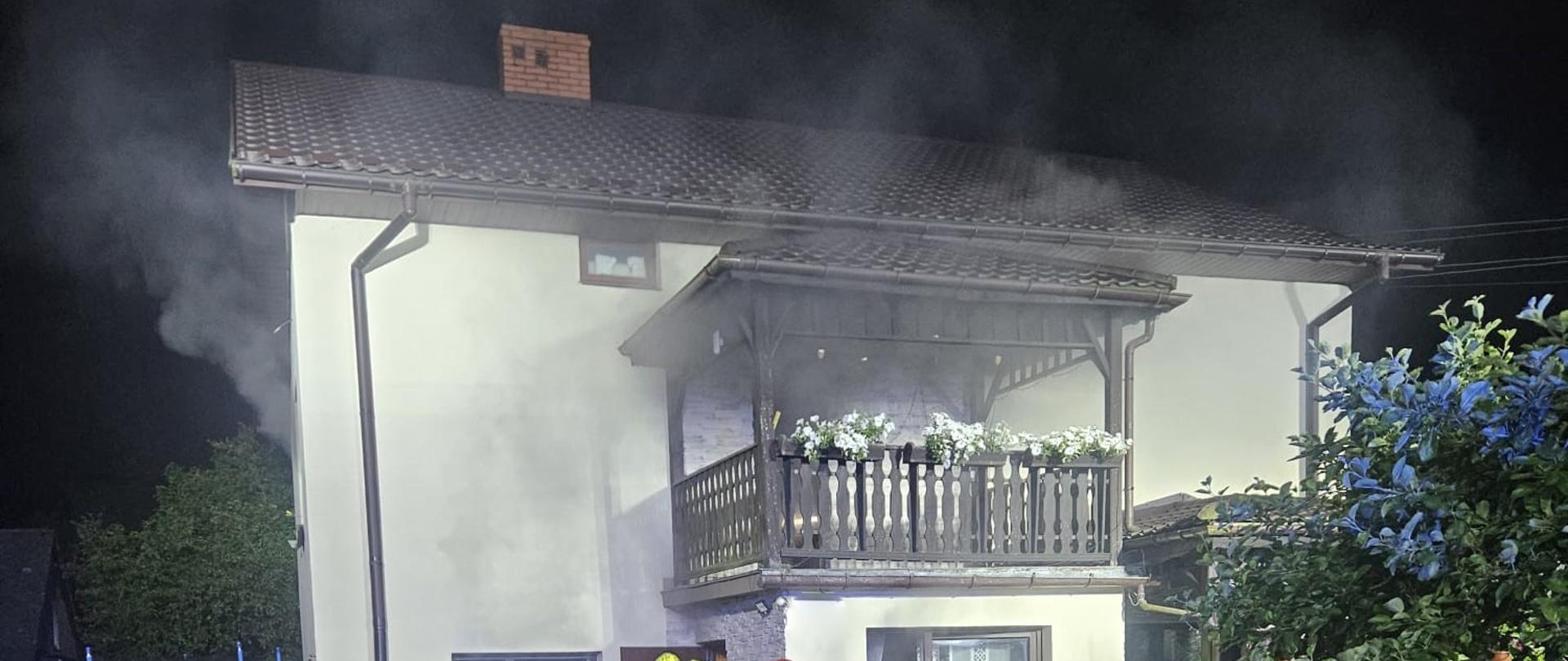 Budynek piętrowy, jednorodzinny, z którego wydobywa się dym. Koło budynku pracujący strażacy.