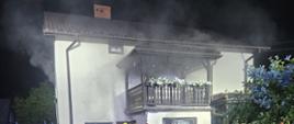 Budynek piętrowy, jednorodzinny, z którego wydobywa się dym. Koło budynku pracujący strażacy.