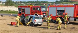 Zdjęcie przedstawia strażaków, którzy prowadzą ćwiczenia z zakresu ratownictwa technicznego. Dwóch strażaków znajduje się w samochodzie osobowym. W tle znajdują się dwa wozy strażackie