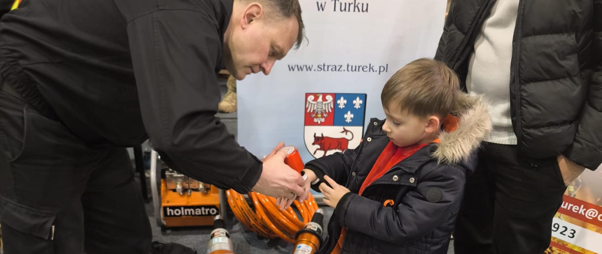 Strażak w czarnym mundurze pokazuje małemu chłopcu sprzęt ratowniczy; dziecko stoi obok dorosłego mężczyzny. Na podłodze leżą hydrauliczne narzędzia strażackie i puzzle z rysunkiem strażaka. W tle baner Komendy Powiatowej Państwowej Straży Pożarnej w Turku oraz sala sportowa.