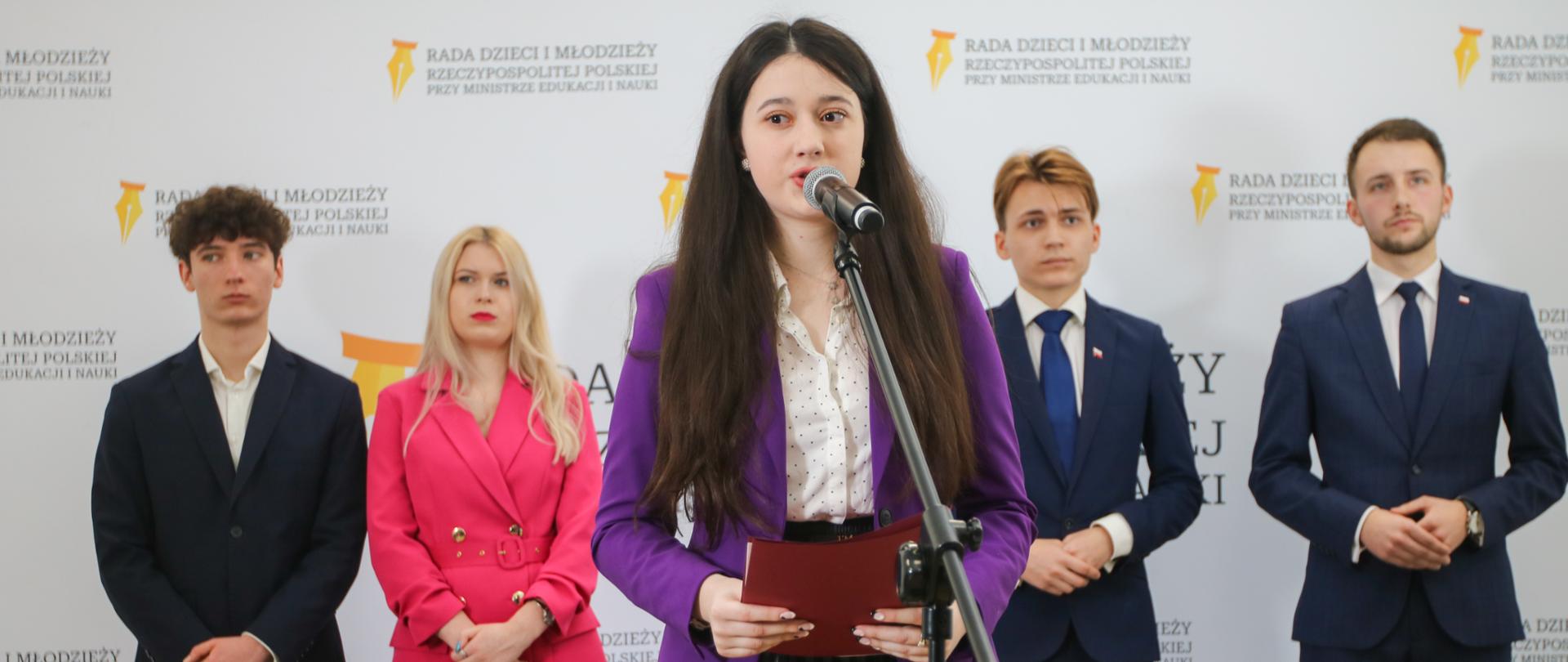 Młoda dziewczyna w fioletowym sweterku stoi przy mikrofonie na stojaku, za nią trzech młodych mężczyzn w garniturach i dziewczyna w czerwonej sukience, za nimi biała ściana z napisem Rada Dzieci i Młodzieży.