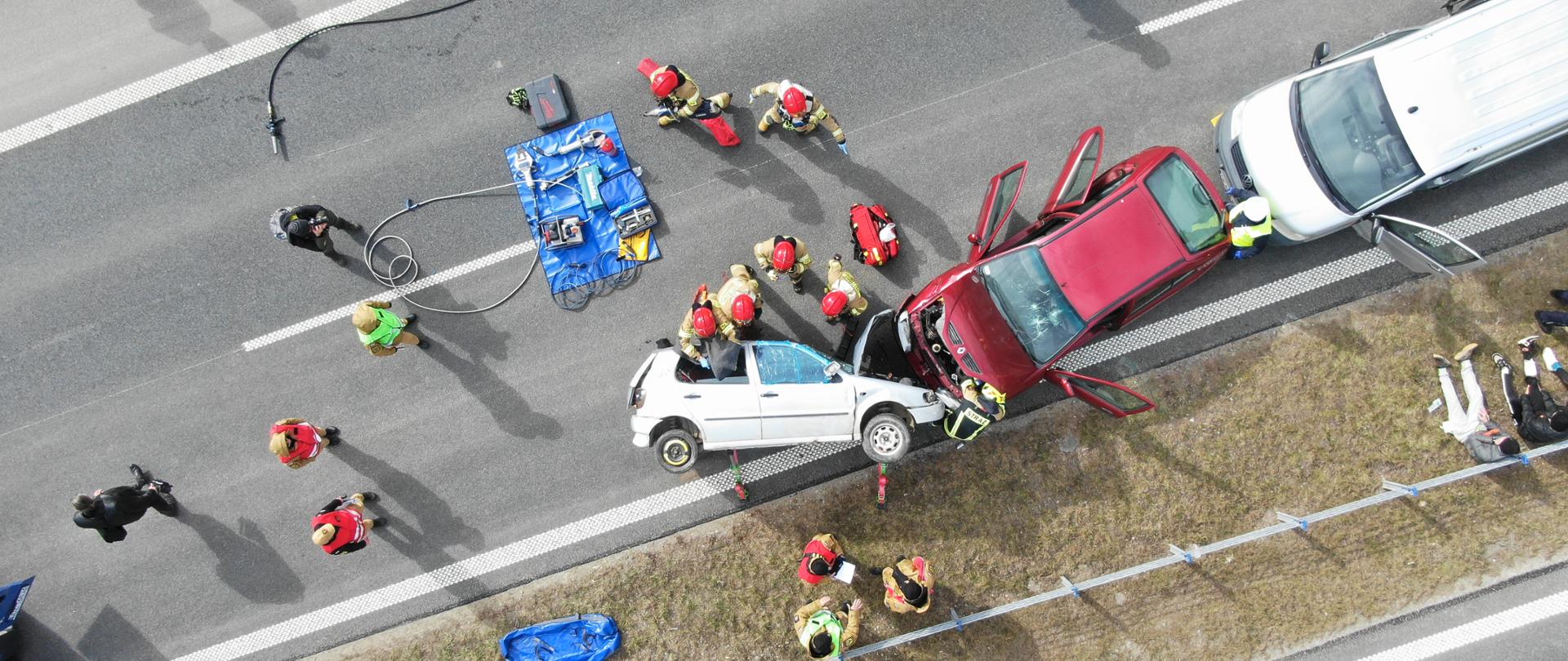 ćwiczenia służb ratowniczych widziane z góry. Na jezdni rozbite w stłuczce samochody, służby ratownicze pomagają poszkodowanym