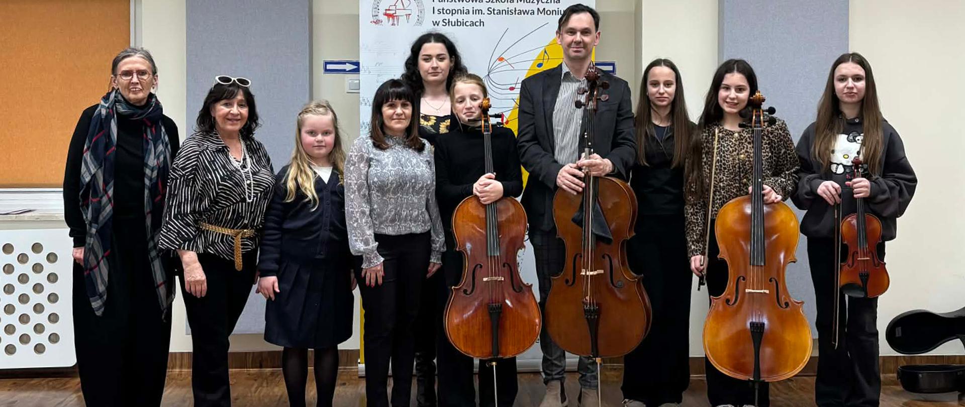 Na zdjęciu stoi grupa kilkunastu osób, niektórzy w rękach trzymają wiolonczele i skrzypce.