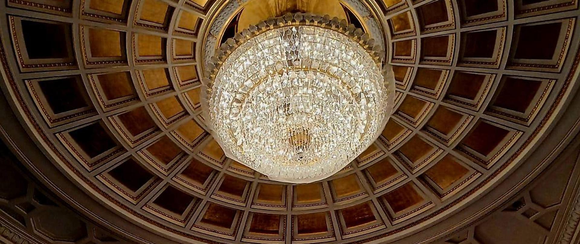 Wnętrze Teatru Wielkiego w Poznaniu. Widok na bogato zdobioną widownię z kilkoma poziomami balkonów, czerwonymi ścianami i złoconymi detalami architektonicznymi. Nad salą dominuje duży kryształowy żyrandol zawieszony pod ozdobną, kasetonową kopułą