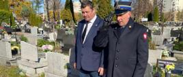 Zdjęcie dokumentuje uroczysty moment oddania hołdu na cmentarzu, w przededniu Dnia Wszystkich Świętych.
Osoby: W centrum kadru znajdują się dwaj mężczyźni.
Mężczyzna po lewej stronie to prawdopodobnie Komendant Miejski Państwowej Straży Pożarnej w Mysłowicach, st. bryg. Rafał Pietrzyk. Ubrany jest w ciemny, galowy lub służbowy mundur PSP, z widocznymi dystynkcjami i odznaczeniami. Komendant stoi tyłem do grobu, wykonując gest szacunku, być może poprawiając wstęgę lub spoglądając na grób.
Drugi mężczyzna, stojący po prawej stronie, ubrany jest w ciemny strój cywilny (prawdopodobnie garnitur lub marynarkę) i pomaga w składaniu wieńca lub wiązanki.
Grób i hołd: Osoby znajdują się przy nagrobku, na którym spoczywają złożone wieńce i wiązanki. Na pierwszym planie widoczne są kwiaty w barwach narodowych (biało-czerwone) lub kolory związane ze strażą.
Ważnym elementem są liczne zapalonych znicze i lampiony, które symbolizują pamięć, szacunek i wdzięczność, zgodnie z tradycją przed dniem Wszystkich Świętych.
Tło: Tło zdjęcia jest charakterystyczne dla cmentarza – widoczne są elementy innych nagrobków, co wzmacnia podniosły i refleksyjny nastrój.
Zdjęcie jest ilustracją do przekazu o pielęgnowaniu pamięci o strażakach, którzy odeszli na wieczną służbę i oddaniu im hołdu przez kierownictwo Komendy Miejskiej PSP w Mysłowicach.