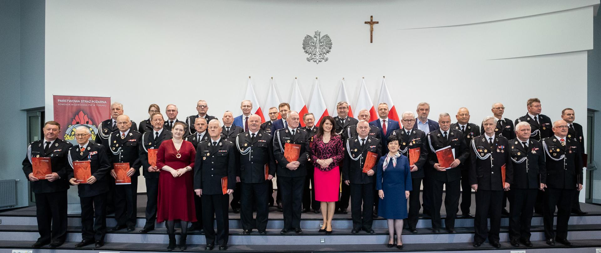 Zdjęcie przedstawia wnętrze obszernego pomieszczenia, w którym na schodach stoi kilkadziesiąt osób. Większość z nich to mężczyźni w galowych ciemnych mundurach, którzy w rękach trzymają czerwone teczki. Pozostali to osoby cywilne kobiety w sukniach i mężczyźnie w garniturach. W tle na ścianie wisi srebrny orzeł w koronie i krzyż.