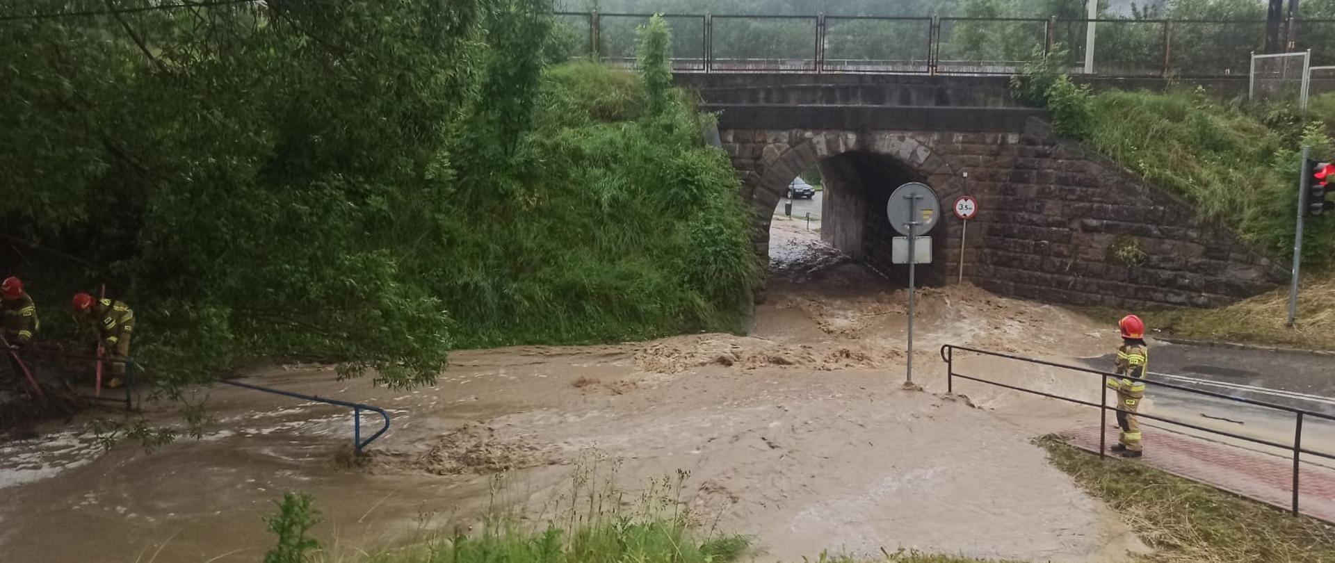 Woda płynie droga a obok strażacy
