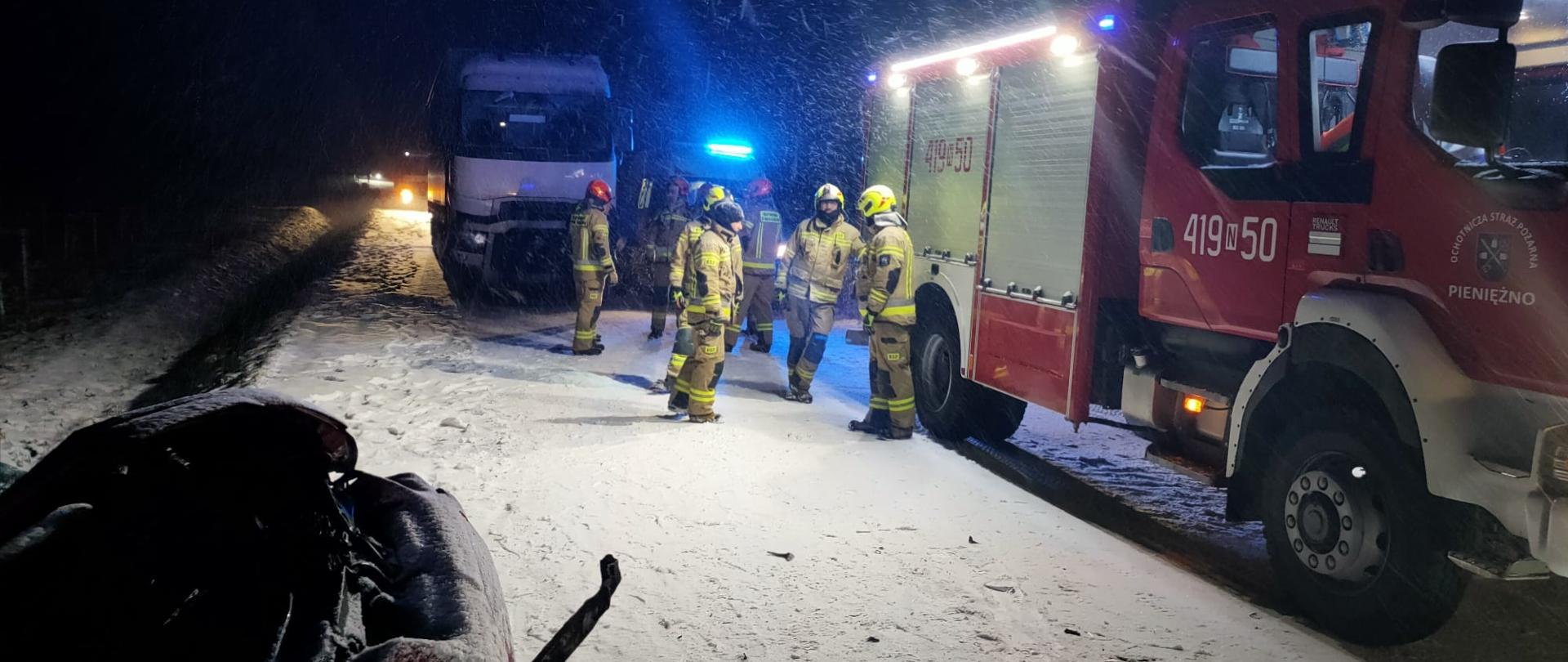 Noc. Na zaśnieżonej drodze z prawej strony stoi duży samochód strażacki. Obok strażacy. Pada śnieg. W dolnym lewym rogu uszkodzone auto w rowie.