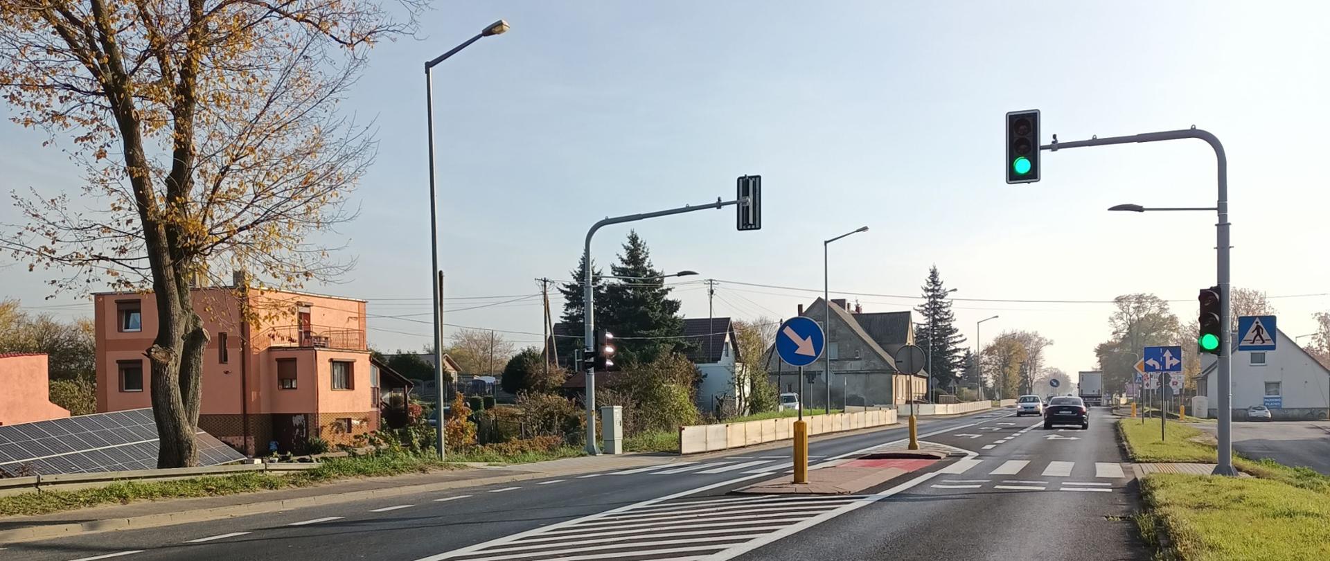 na drodze przebiegającej przez miejscowość widoczne jest przejście dla pieszych wraz z sygnalizacją świetlną. w tle widoczne zabudowania po dwóch stronach drogi. centralnie na zdjęciu widoczne są poruszające się samochody.