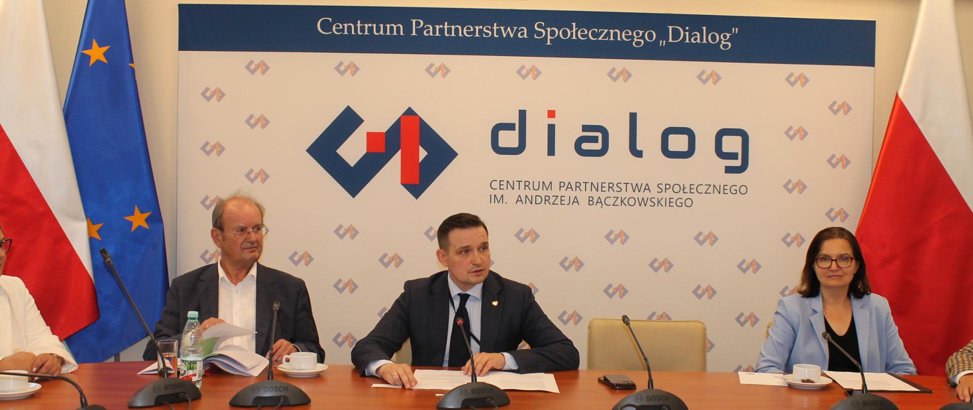 Na zdjęciu widnieje stół prezydialny za którym siedzą: Pan Tadeusz Wawrzyniak, Współprzewodniczący Zespołu ze strony pracodawców; Pan Michał Jaros, Sekretarz Stanu w Ministerstwie Rozwoju i Technologii oraz Pani Anita Sowińska, Podsekretarz Stanu w Ministerstwie Klimatu i Środowiska. W tle znajduje się baner z napisem Centrum Partnerstwa Społecznego „Dialog”. Po lewej stronie znajdują się dwie flagi – Polski oraz Unii Europejskiej. Po prawej stronie znajduje się flaga Polski. Nad banerem widnieje Godło Polski.