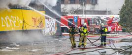 Zdjęcie przedstawia pożar budynku usługowego oraz akcję gaśniczą. Na zdjęciu widoczni czterej strażacy podający dwa prądy wody do wnętrza obiektu (w kierunku pożaru). W tle widoczny czerwone samochody straży pożarnej.