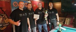 Czterech mężczyzn stoi razem, pozując do zdjęcia po zawodach, na tle banerów związanych ze Strażą Pożarną w Wałbrzychu. Wszyscy ubrani są w czarne koszulki polo, prawdopodobnie jednakowe, z napisem "NSZZP" (Niezależny Samorządny Związek Zawodowy Policjantów) lub podobnym logo na piersi. Mężczyzna po lewej trzyma kciuk w górze. Dwóch mężczyzn w środku trzyma nagrody – jeden trzyma białą kopertę lub kartkę, a drugi – większy złoty puchar. Przed nimi, na skraju stołu bilardowego, stoi mniejszy zielono-złoty puchar. W tle widać dekoracje na suficie i czerwone banery