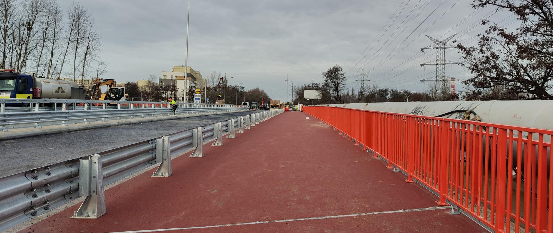 Widok z przebudowanego wiaduktu w ciągu ulicy Bohaterów Monte Cassino nad drogą ekspresową S86 w Katowicach. Na pierwszym planie znajduje się szeroka, świeżo wykonana nawierzchnia w kolorze czerwonym, przeznaczona dla pieszych lub rowerzystów, oddzielona od jezdni stalową barierą energochłonną. Po lewej stronie widoczna jest jezdnia z nową warstwą asfaltu, na której stoją maszyny budowlane i pracownicy w odzieży roboczej. Po prawej stronie biegnie rząd pomarańczowych barier zabezpieczających, a w tle widać słupy wysokiego napięcia, drzewa bez liści oraz zabudowania miejskie. 