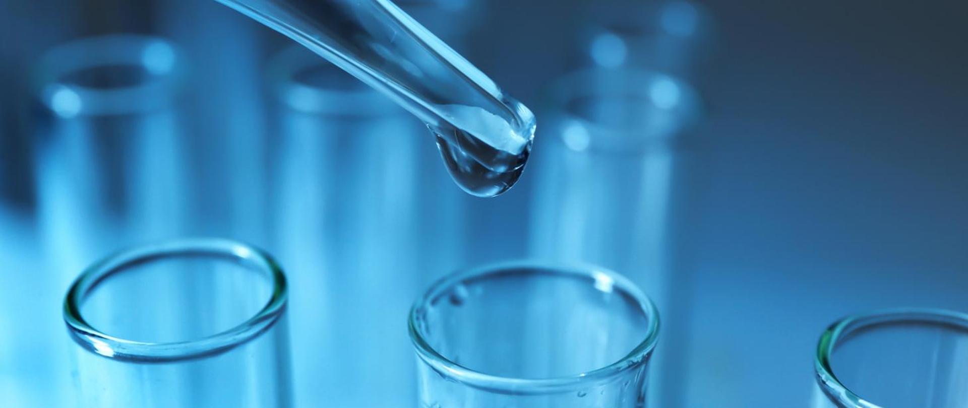 Zbliżenie na szklaną pipetę, z której kropla przezroczystego płynu wpada do probówki w laboratorium.