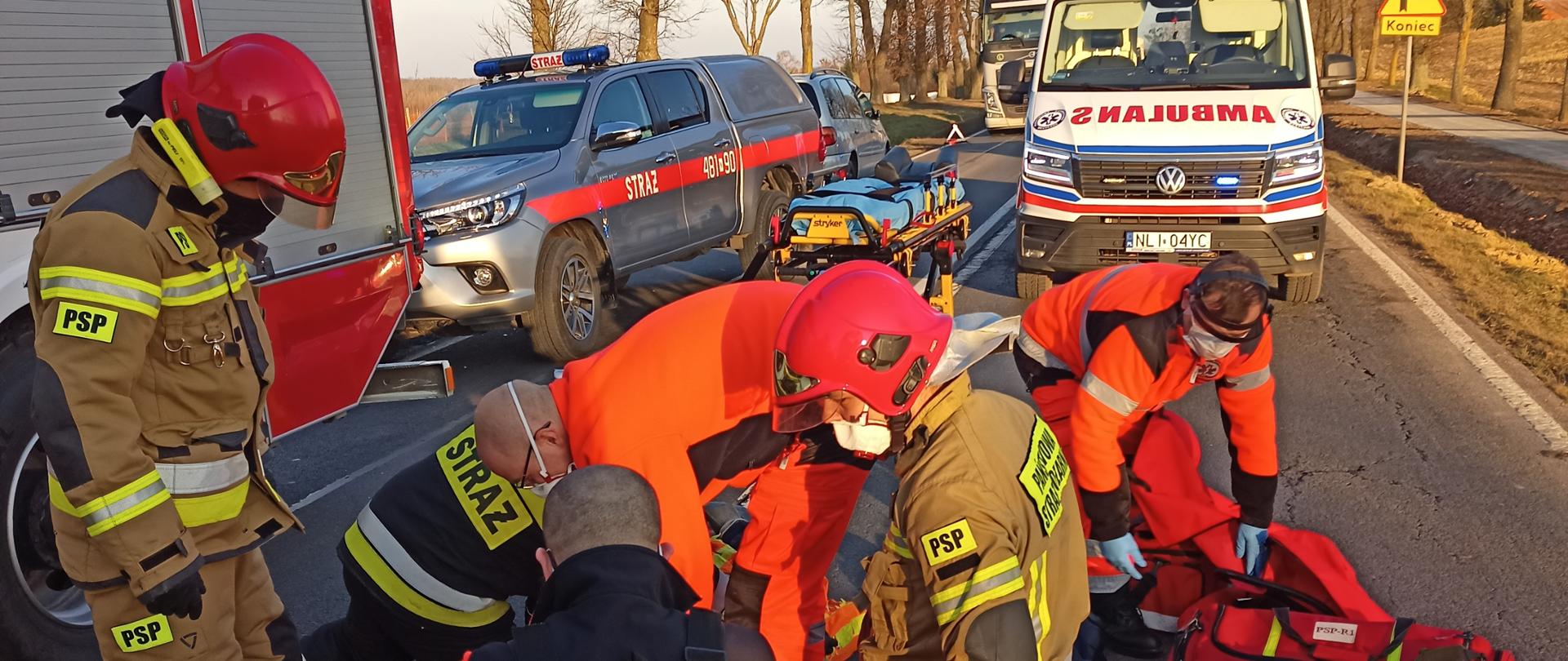Na zdjęciu widać współprace służb ratowniczych. Trzech strażaków PSP oraz trzech medyków, którzy zaopatrują wstępnie osobę poszkodowaną. Poszkodowany leży na asfalcie, został potrącony przez samochód osobowy. Na drugim planie znajdują się samochody pożarnicze oraz karetka pogotowia. 