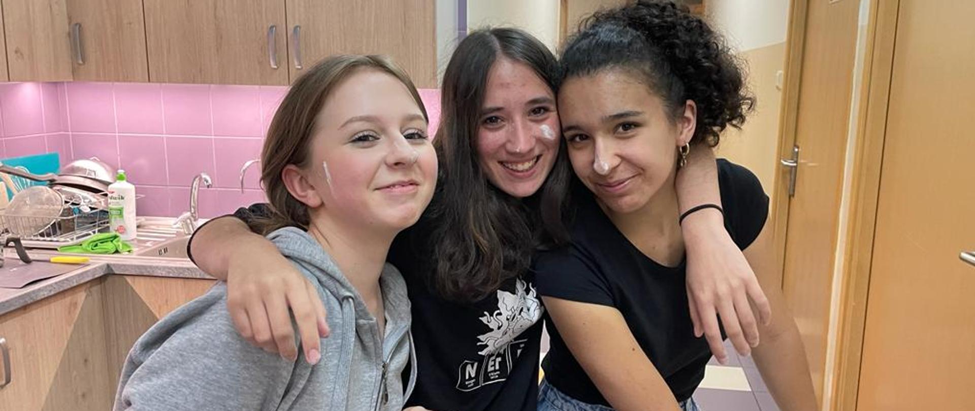 Trzy uśmiechnięte nastolatki przygotowują ciasto na drożdżówki w aneksie kuchennym w internacie.