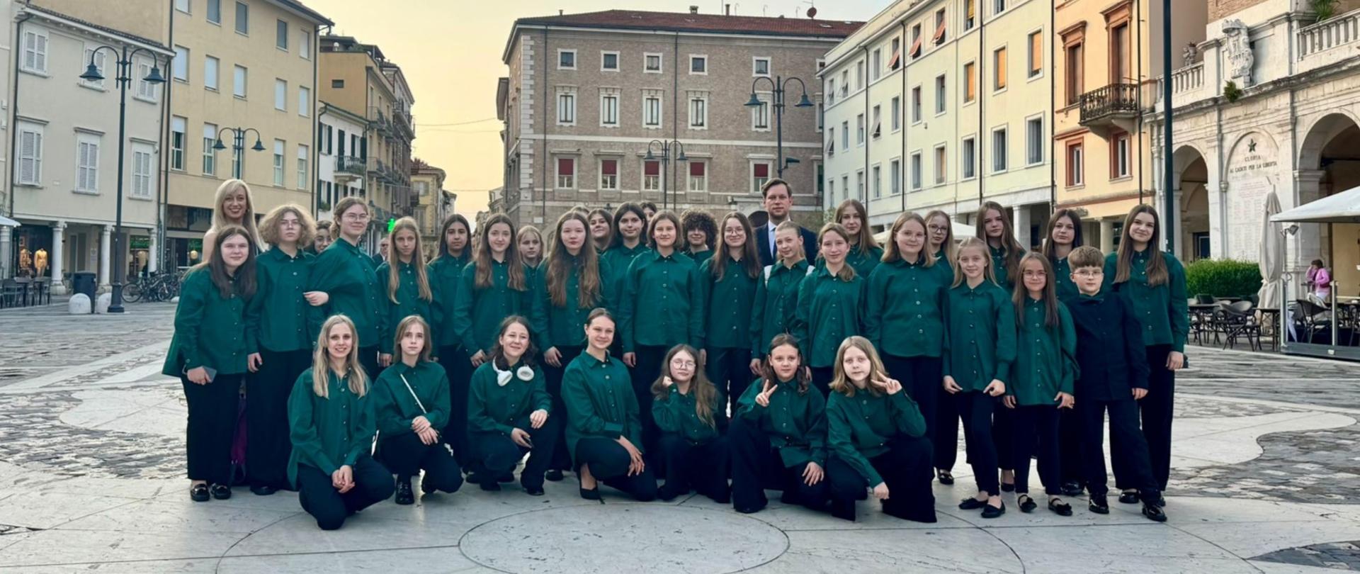 Grupowe zdjęcie chóru młodzieżowego w zielonych koszulach, wykonane na tle włoskiego placu miejskiego otoczonego klasyczną architekturą. W tle widoczne są budynki z pastelowymi fasadami, wieża zegarowa oraz kawiarniane ogródki. Zdjęcie zostało zrobione o zmierzchu, w miękkim świetle zachodzącego słońca.