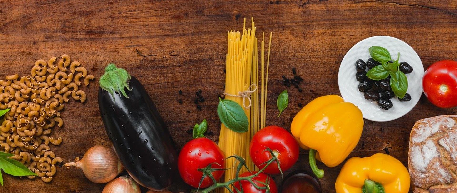 makaron, cebula, bakłażan, pomidory, żółta papryka, ziemne oliwki i chleb leżące na ciemnobrązowym drewnianym blacie