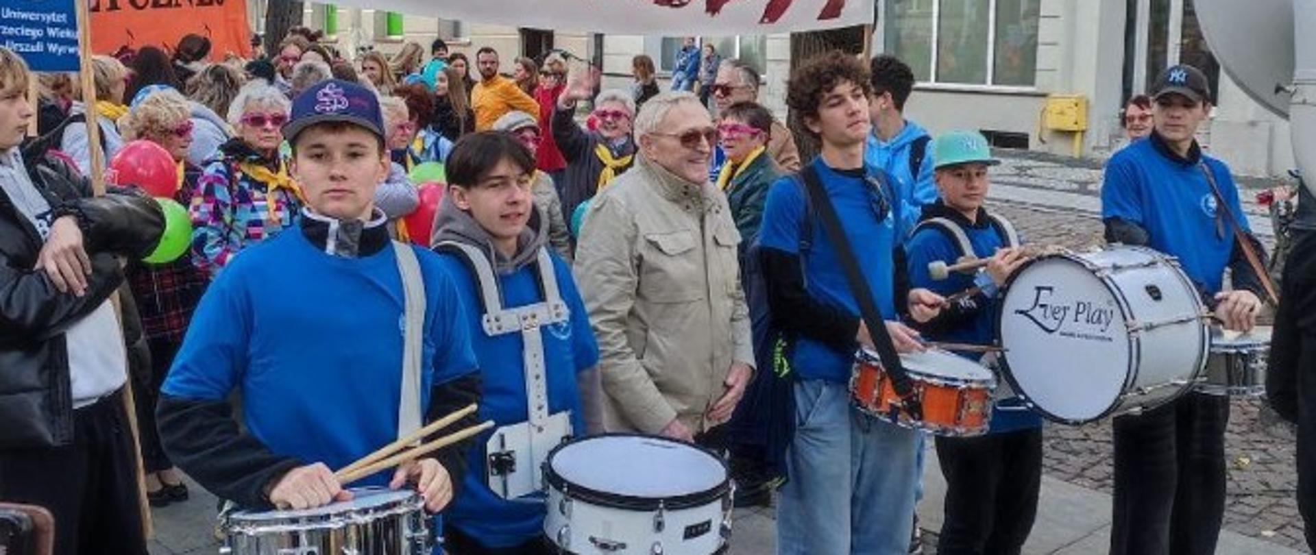 Zdjęcie kolorowe: Grupa osób na ulicy, (prawdopodobnie) podczas parady, jesienią; na pierwszym planie od lewej cztery osoby w niebieskich koszulkach z pałeczkami, grające na werblach; w centrum starszy mężczyzna w beżowej kurtce i ciemnych okularach; nad głowami grupy duży transparent z napisem "KMEDA" w kolorach czerwono-pomarańczowych; tło stanowiące drzewa z żółtymi liśćmi i tłum ludzi.