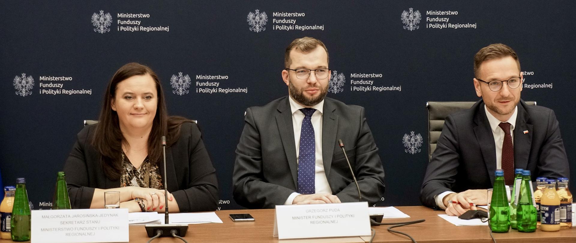Od lewej wiceminister Małgorzata Jarosińska-Jedynak, minister Grzegorz Puda i wiceminister Waldemar Buda siedzą przy stole konferencyjnym. Za nimi ścianka Ministerstwa Funduszy i Polityki Regionalnej.