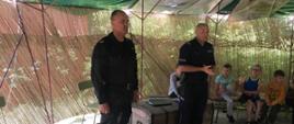 Na zdjęciu wizytacja obozu harcerskiego przez służby mundurowe. Strażak i policjant przekazuje informacje o zagrożeniach na obozowisku. Harcerze siedzą w namiocie i słuchają informacji przekazanych przez służby mundurowe.