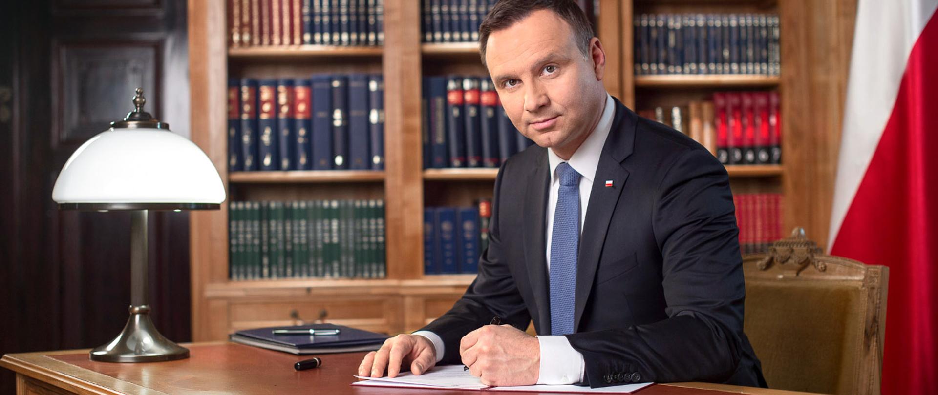 Prezydent Andrzej Duda na tle biało-czerwonej flagi podpisuje dokument