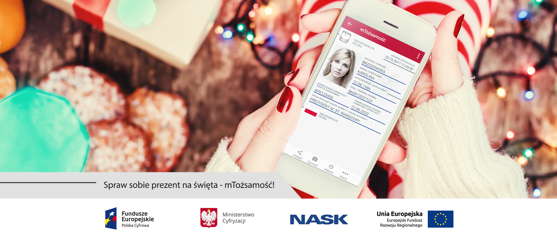 Na pierwszym planie kobiece dłonie trzymające telefon z aplikacją mObywatel, tło świąteczne - niewyraźne lampki, prezenty