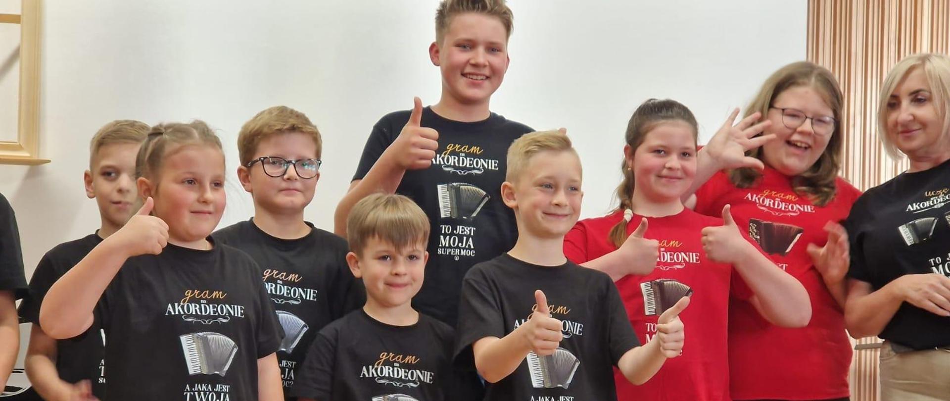 na zdjęciu stoi grupa uczniów z nauczycielką w koszulkach czarnych lub czerwonych z akordeonem