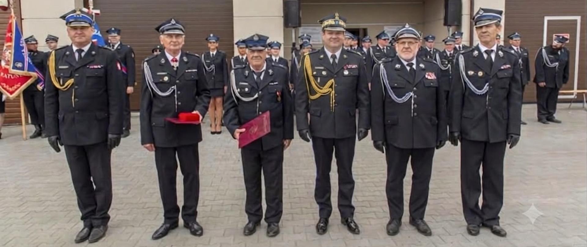 Na zdjęciu przedstawiono grupę strażaków pozujących do pamiątkowej fotografii przed budynkiem remizy Ochotniczej Straży Pożarnej. W pierwszym rzędzie stoją strażacy w galowych mundurach OSP oraz Państwowej Straży Pożarnej, część z nich trzyma czerwone poduszki z odznaczeniami lub pamiątkowymi elementami uroczystości. Za nimi ustawieni są pozostali druhowie i uczestnicy wydarzenia. W tle widoczny jest budynek remizy z otwartymi bramami garażowymi oraz zachmurzone niebo, charakterystyczne dla pogodnego, choć pochmurnego dnia.