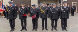 Na zdjęciu przedstawiono grupę strażaków pozujących do pamiątkowej fotografii przed budynkiem remizy Ochotniczej Straży Pożarnej. W pierwszym rzędzie stoją strażacy w galowych mundurach OSP oraz Państwowej Straży Pożarnej, część z nich trzyma czerwone poduszki z odznaczeniami lub pamiątkowymi elementami uroczystości. Za nimi ustawieni są pozostali druhowie i uczestnicy wydarzenia. W tle widoczny jest budynek remizy z otwartymi bramami garażowymi oraz zachmurzone niebo, charakterystyczne dla pogodnego, choć pochmurnego dnia.