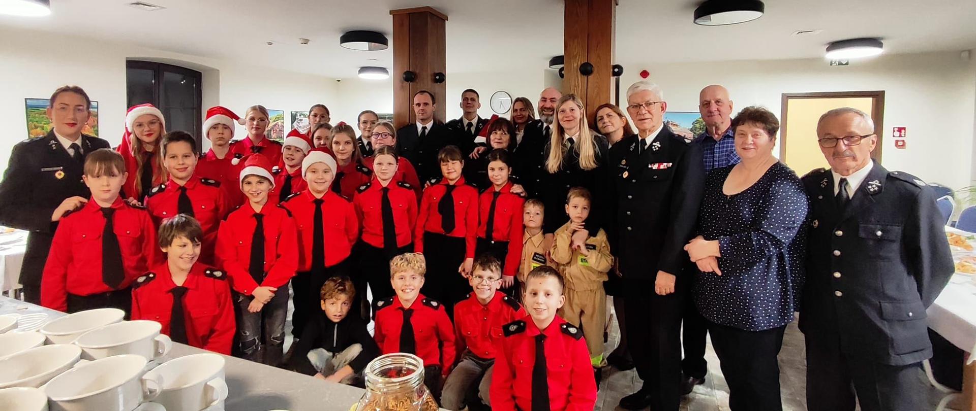 Na zdjęciu duża grupa grupa strażaków ochotników oraz dzieci z młodzieżowej drużyny pożarniczej w czerwonych mundurkach.