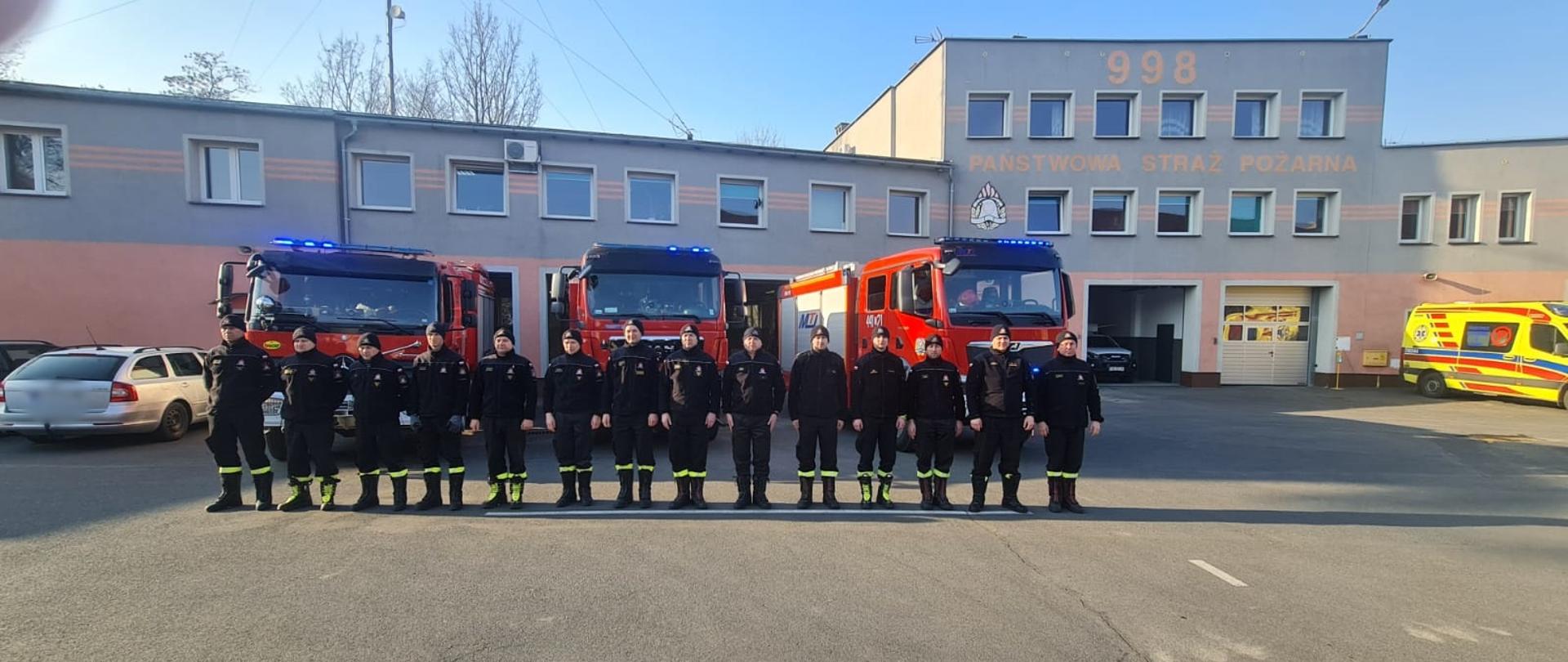 Obraz przedstawia strażaków JRG Jawor podczas zmiany służby podczas której minutą ciszy uczczono pamięć poległych strażaków z Ukrainy. Strażacy stoją w szeregu. W tle samochody pożarnicze i budynek komendy. Pora dzienna.