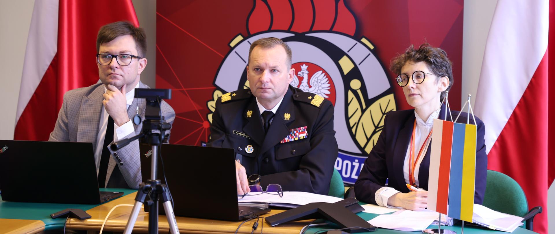 Od lewej: mężczyzna w garniturze, zastępca komendanta w mundurze generalskim oraz kobieta siedzący przy stoliku. W tle flagi polski oraz baner Państwowej Straży Pożarnej. Z boku z prawej strony na stole flagi Polski i Ukrainy