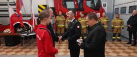 Komendanta Powiatowego wraz z Burmistrzem Gostynia wręczających kluczyki do nowego samochodu druhowi z Ochotniczej Straży Pożarnej w Goli. Obok druha stoi dziewczyna ubrana w czerwony dres Młodzieżowej Drużyny Pożarniczej. Z tyłu stoją druhowie w szeregu przed nowym wozem strażackim.
