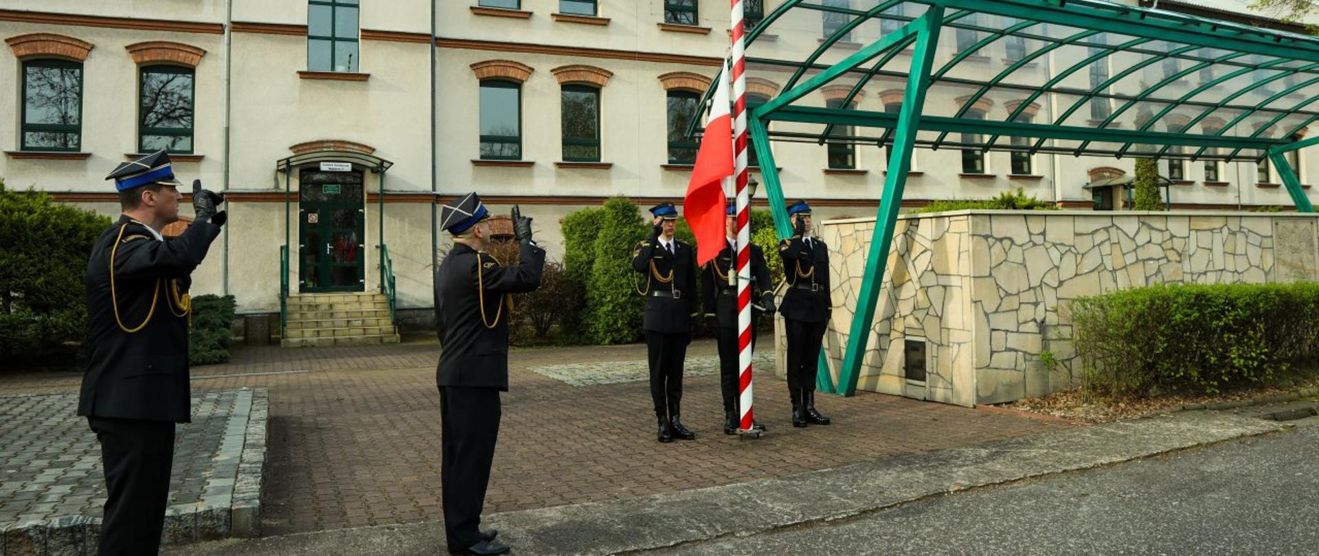 Komendant Centralnej Szkoły PSP oraz dowódca uroczystości salutują podczas gdy na masz wciągana jest biało-czerwona flaga. U podnóża masztu trzech strażaków pocztu sztandarowego w mundurach galowych. W tle budynek dydaktyczny Szkoły