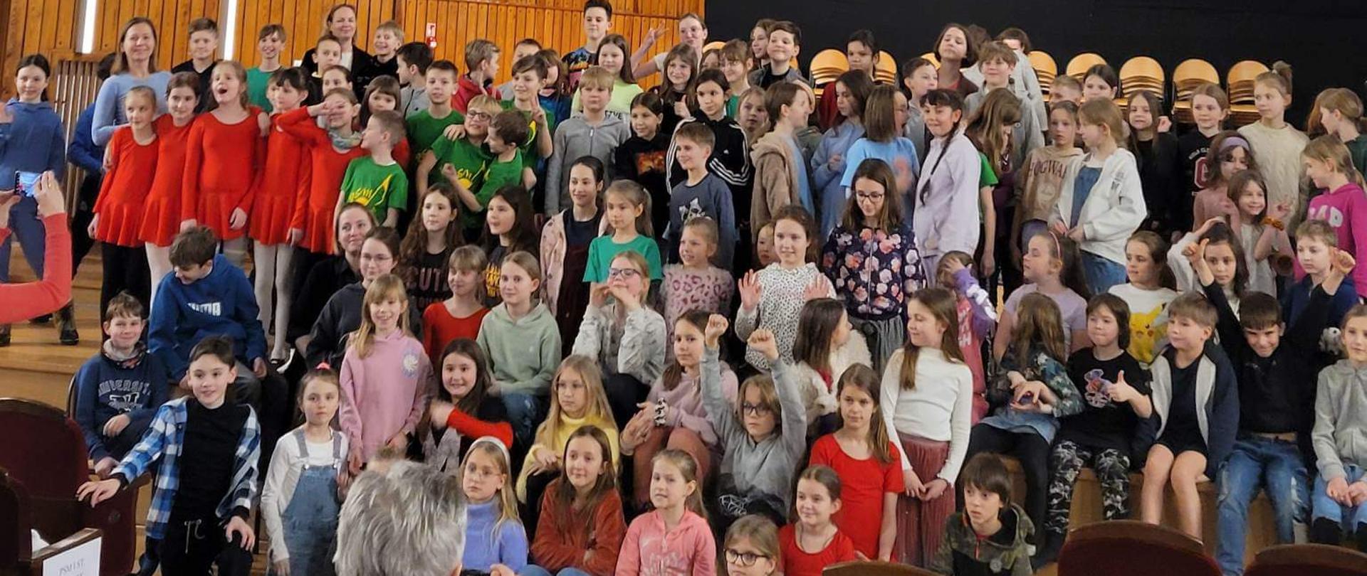 Fotografia przedstawia dużą grupę uczniów siedzących na scenie podczas Warsztatów umuzykalniających