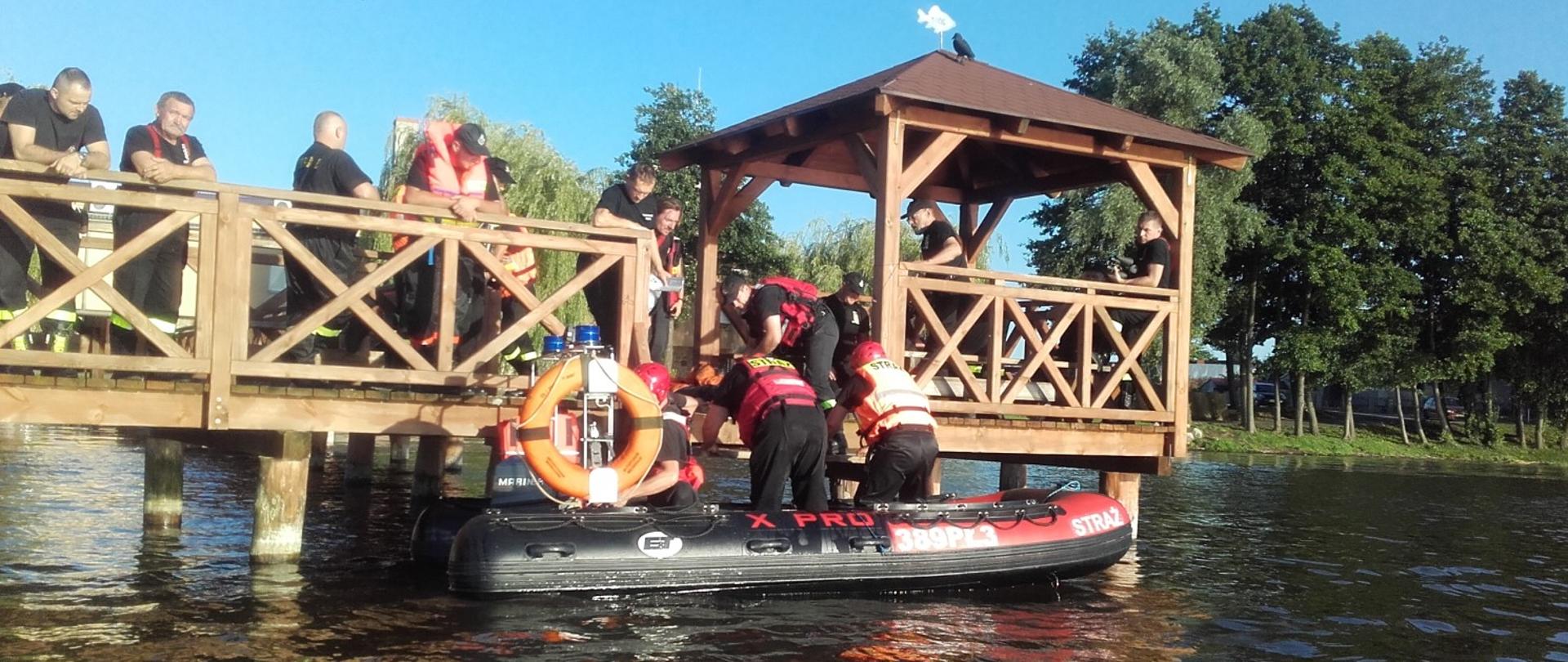 Na zdjęciu widoczni strażacy w pontonie ratowniczym opuszczonym na wodzie przygotowujący się do działań oraz na pomości drewnianym. 