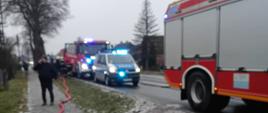 Zabezpieczony odcinek drogi podczas akcji gaśniczej – wozy straży pożarnej i Policji z włączonymi sygnałami świetlnymi oraz wąż gaśniczy ułożony wzdłuż jezdni.